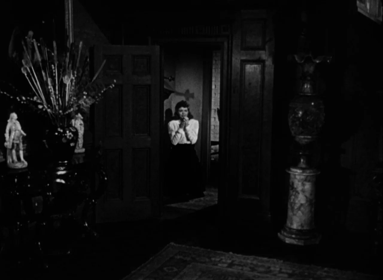 The Spiral Staircase (1946) movie still: medium — A dark hallway in a mansion features ornate furniture in the foreground. A woman stands…