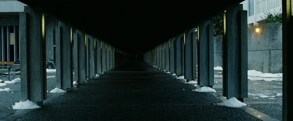 Wind Chill (2007) movie still: establishing — A long, symmetrical covered walkway is lined with rhythmic concrete pillars under a dark sky. Patches…