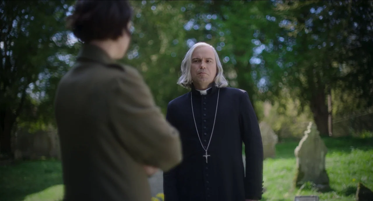 Men (2022) movie still: medium — A rural graveyard with weathered stone monuments surrounded by dense, leafy trees. A priest stands facing another…