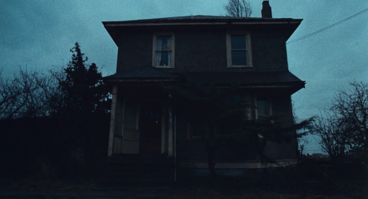Out of the Blue (1980) movie still: establishing — A two-story residential building with dark siding stands against a dim blue sky. The structure…