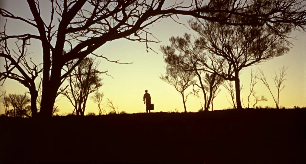 Wake In Fright (1971) movie still: wide — A silhouette of a person walks across a barren ridge at sunset. Bare, spindly trees frame the subject…