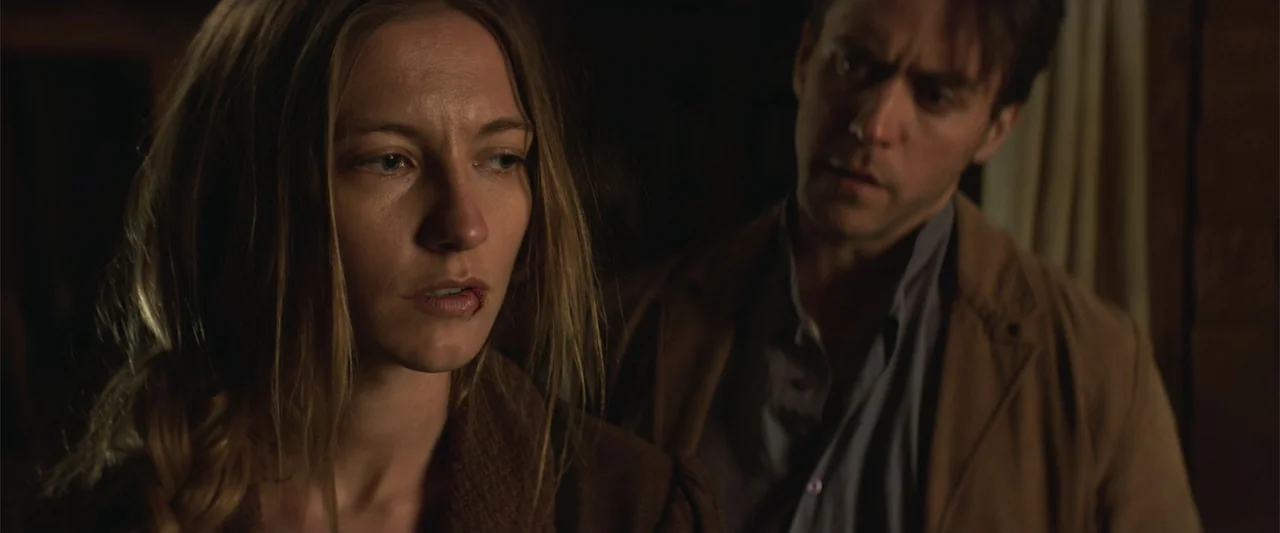 The Wind (2005) movie still: medium — A dimly lit interior space with dark walls and minimal environmental detail visible. A woman with an injured…