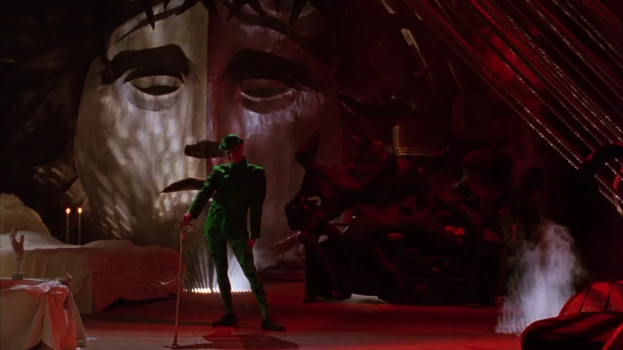 Batman Forever (1995) movie still: wide — A dark subterranean chamber features a massive sculptural mask of a face with thorns. The Riddler stands…