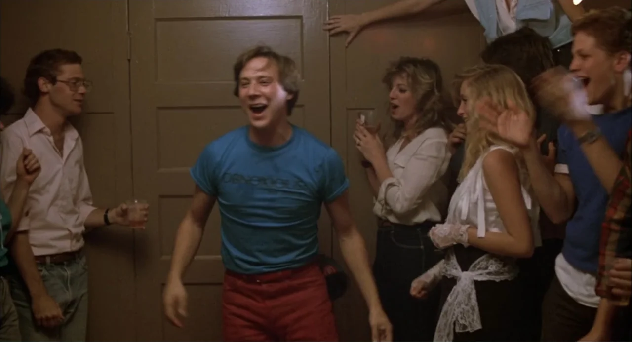 Teen Wolf (1985) movie still: medium — A crowded indoor party room features a plain door as a background backdrop. Several young adults stand…