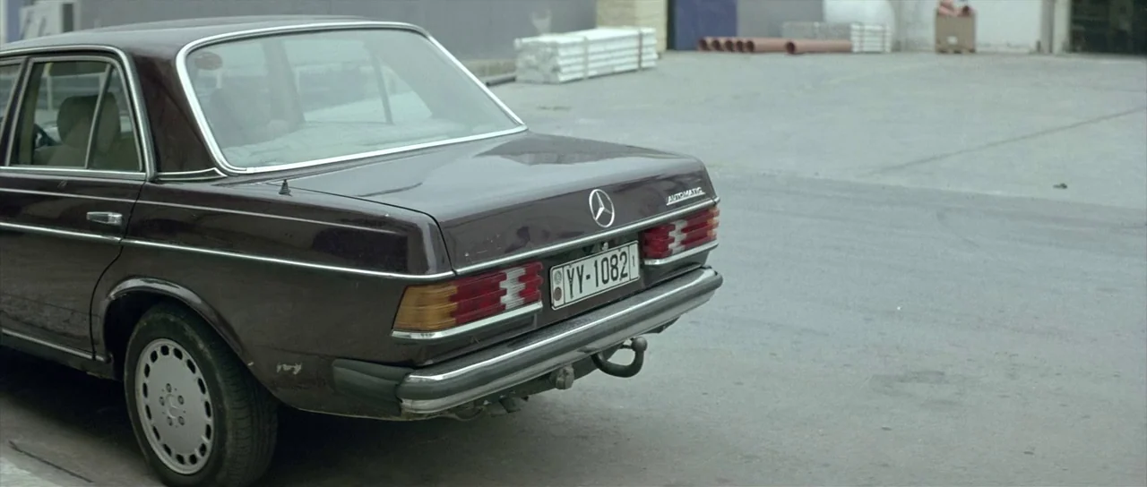 Dogtooth movie still: medium — A dark purple vintage Mercedes-Benz sedan sits parked on a grey asphalt lot. Concrete pipes and building materials…