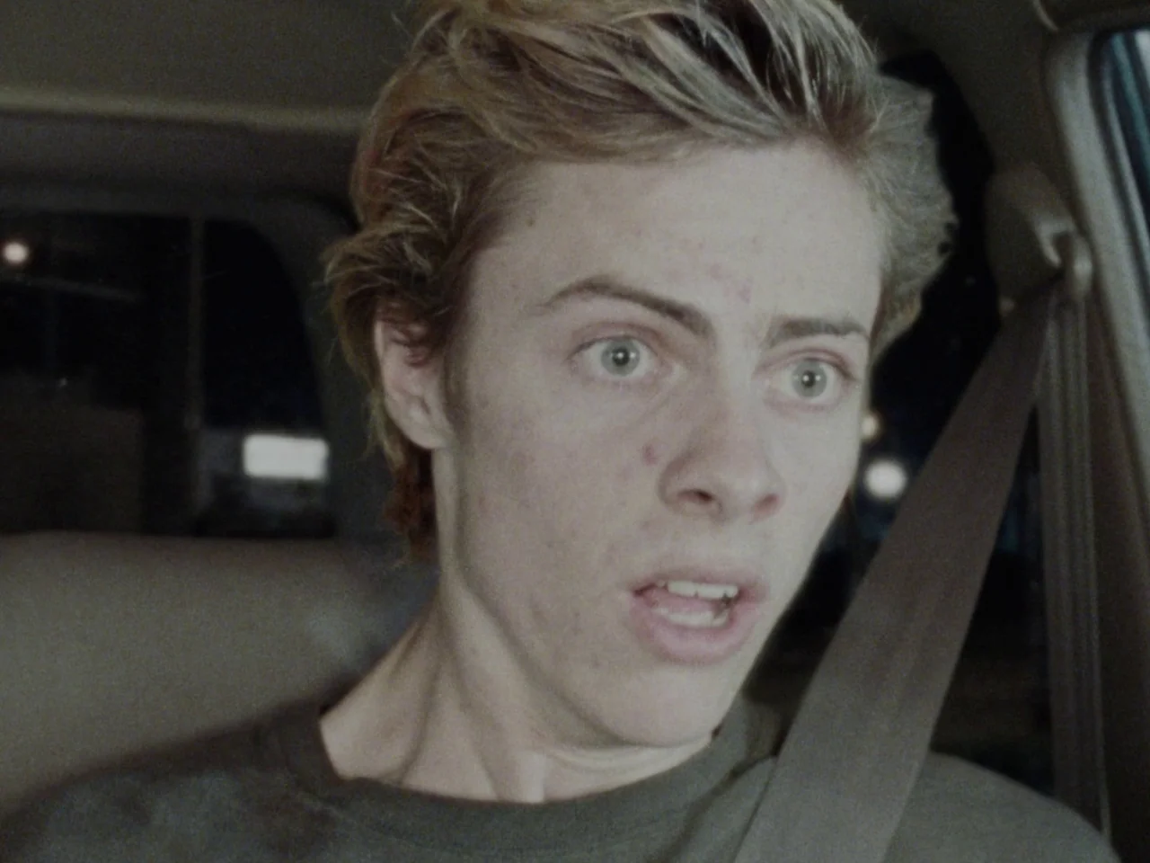 Mid90s (2018) movie still: closeup — A dark vehicle interior with the passenger seat and seatbelt clearly visible. A young man sits in the passenger…