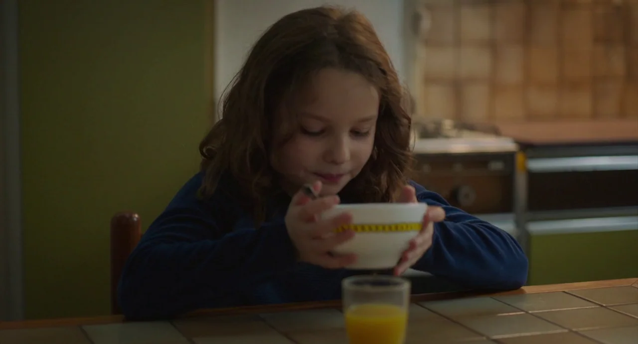 Petite Maman (2021) movie still: medium — A child sits at a tiled dining table with a bowl and glass. She holds the white bowl steady with both…