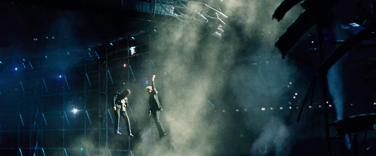 The Island (2005) movie still: wide — A vast, dark arena interior filled with light haze and metallic scaffolding structures. Two men hang suspended…