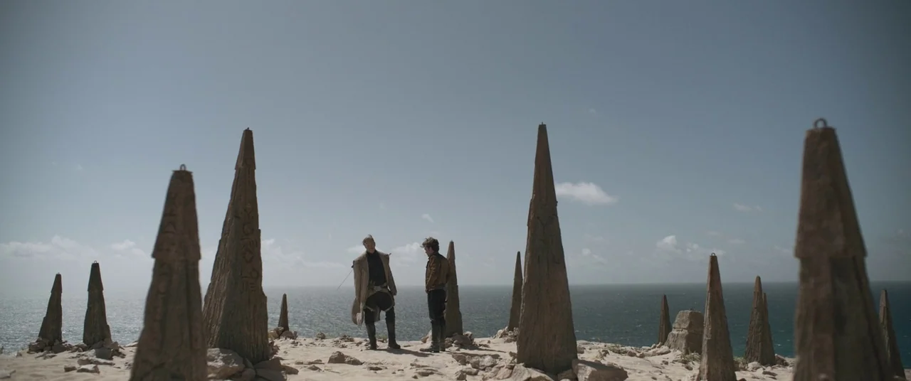 Solo: A Star Wars Story (2018) movie still: wide — A desolate clifftop overlooking the ocean features scattered, tall, weathered stone obelisks. Two…