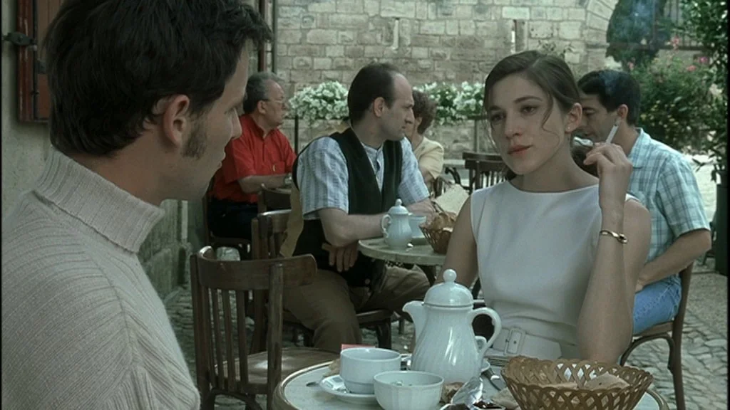 Romance (1999) movie still: medium — An outdoor cafe terrace with cobblestone flooring and stone walls. A man and a woman sit at a round table with…