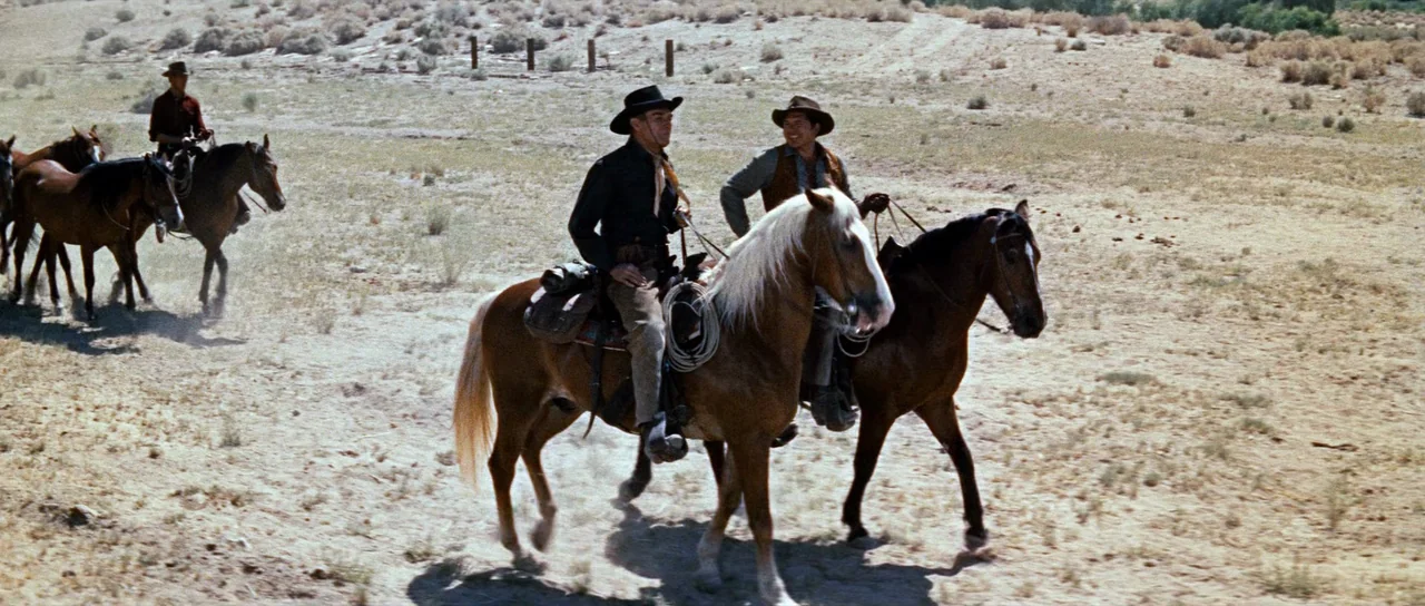 Comanche Station (1960) movie still: wide — A dry, sandy desert landscape with sparse scrub brush and dry grasses. Three cowboys on horseback ride…