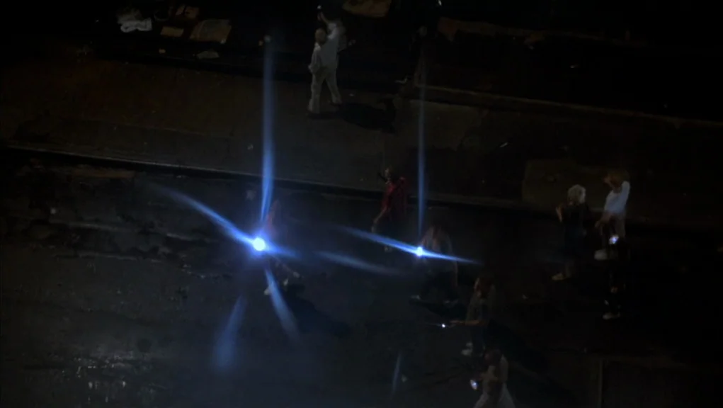 After Hours (1985) movie still: wide — A dark, empty city street at night is illuminated by bright streetlamp light sources. Several figures walk…