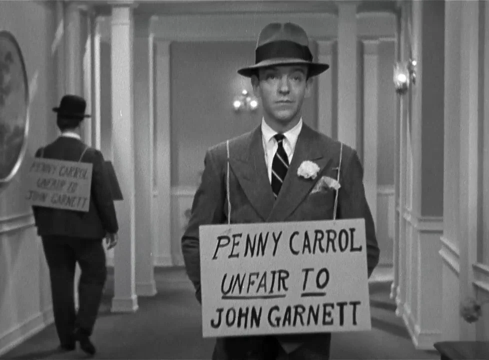 Swing Time (1936) movie still: medium — An interior hotel hallway with white columns and wallpaper is lit by overhead lamps. A man in a suit wears a…