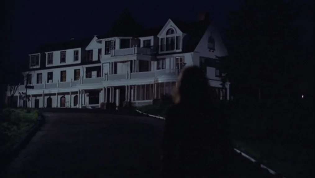 Sisters (1973) movie still: wide — A large multi-story Victorian-style mansion stands against a dark, night-time sky. A person walks along a paved…