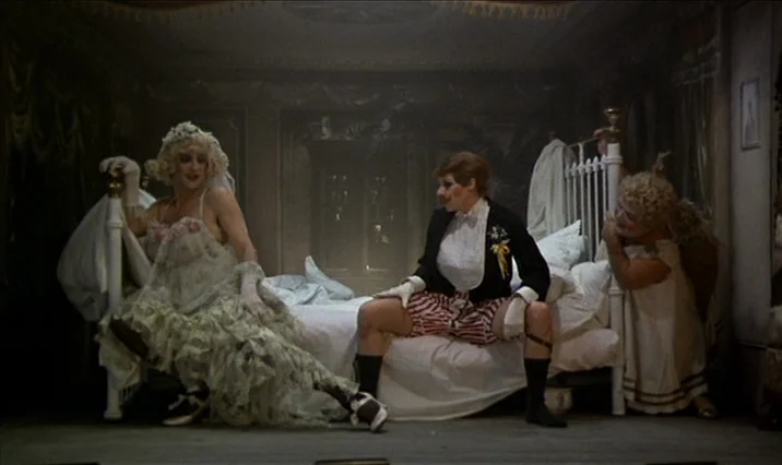 The Serpent’s Egg (1977) movie still: fullbody — A dimly lit room contains a white iron-frame bed in the center area. Three figures in stylized,…