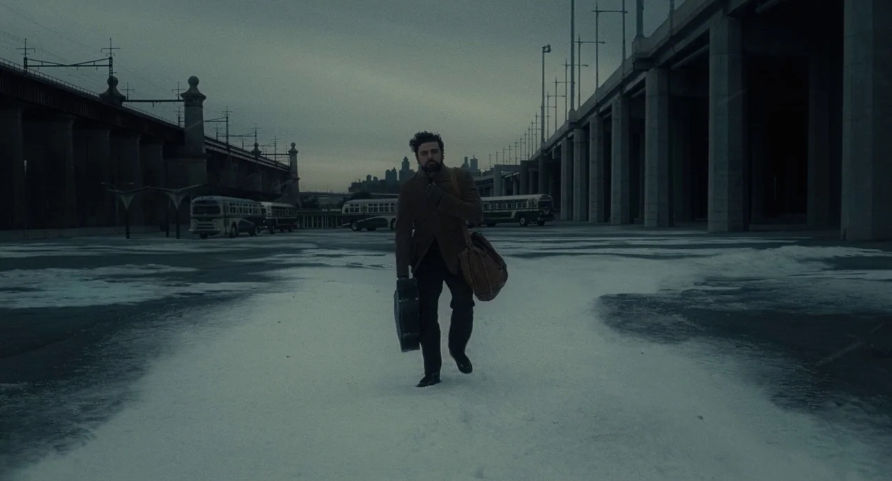 Inside Llewyn Davis (2013) movie still: fullbody — A snowy, concrete-paved bus depot is surrounded by industrial overpasses under a grey sky. A man…