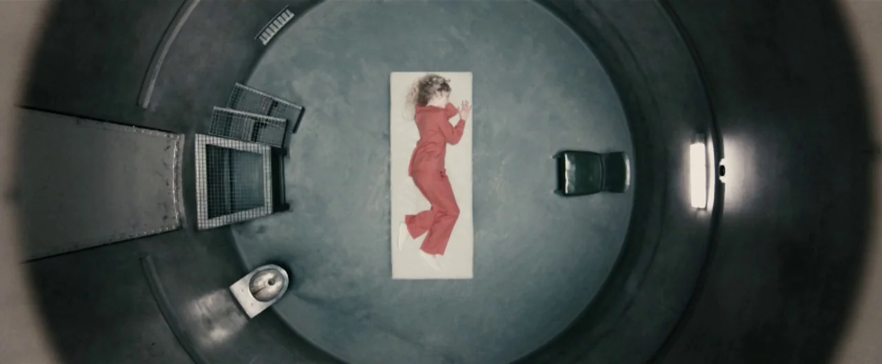 Hanna (2011) movie still: wide — A circular concrete confinement space contains a small mattress, a metal stool, and a toilet. A lone figure in a…