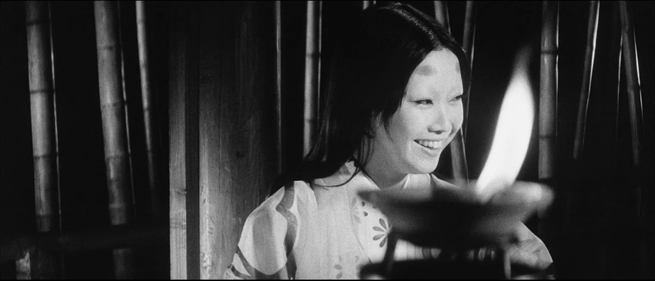 Kuroneko (1968) movie still: medium — A woman sits in a dimly lit, dark space backed by vertical wooden beams. She beams toward a light source…