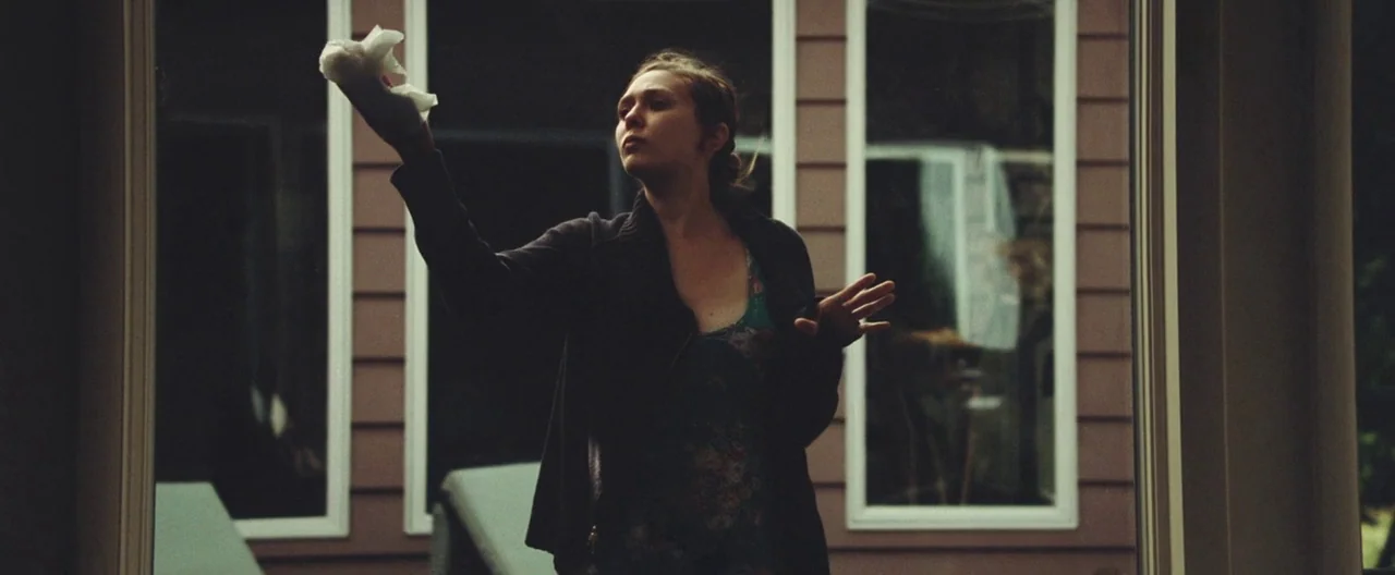 Martha Marcy May Marlene (2011) movie still: medium — A woman stands before a large glass window pane set in a wood frame. She uses a crumpled white…