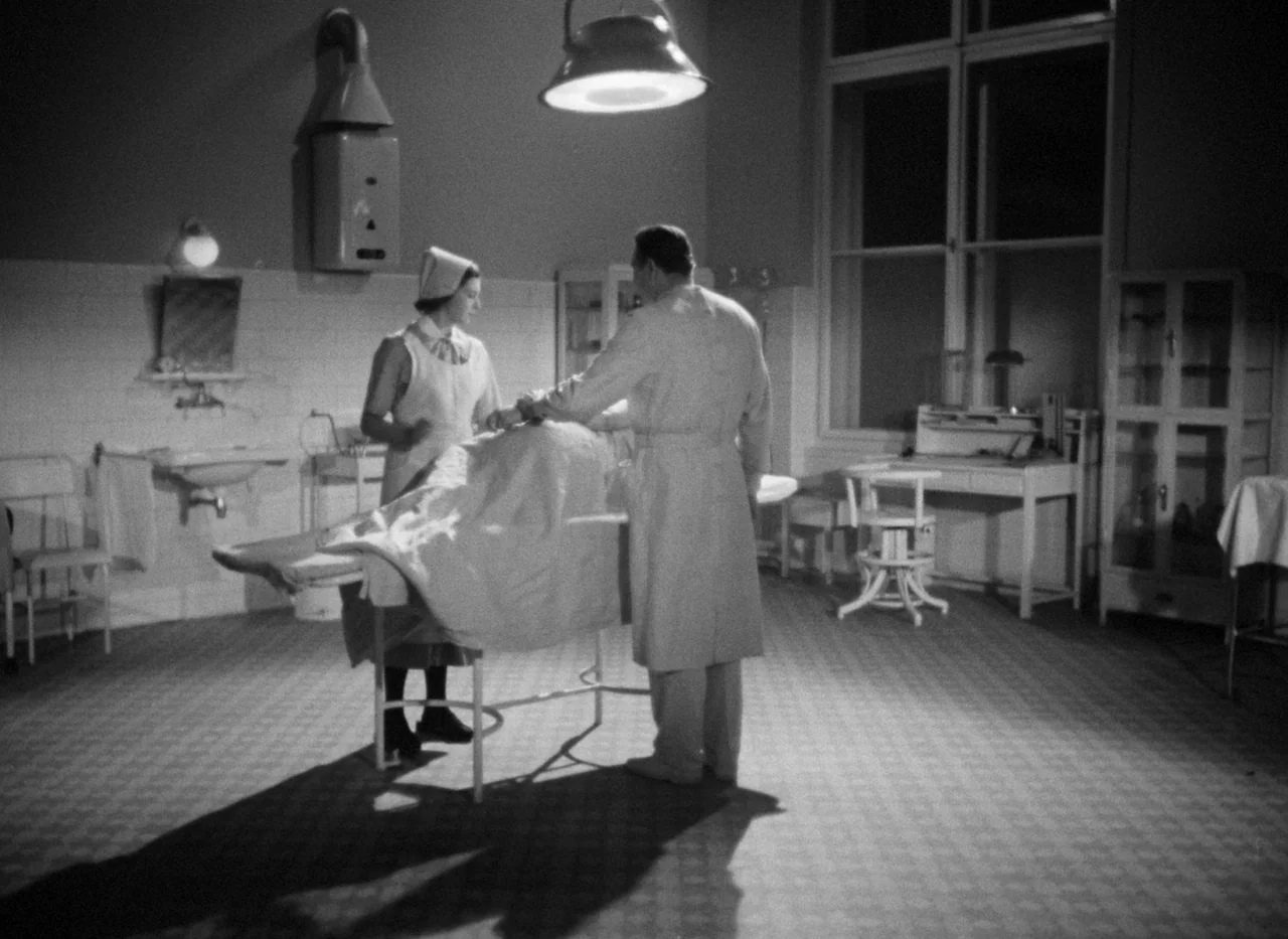 Krakatit (1948) movie still: fullbody — A spartan 1940s medical room contains a central metal examination table and tiled flooring. A doctor and a…