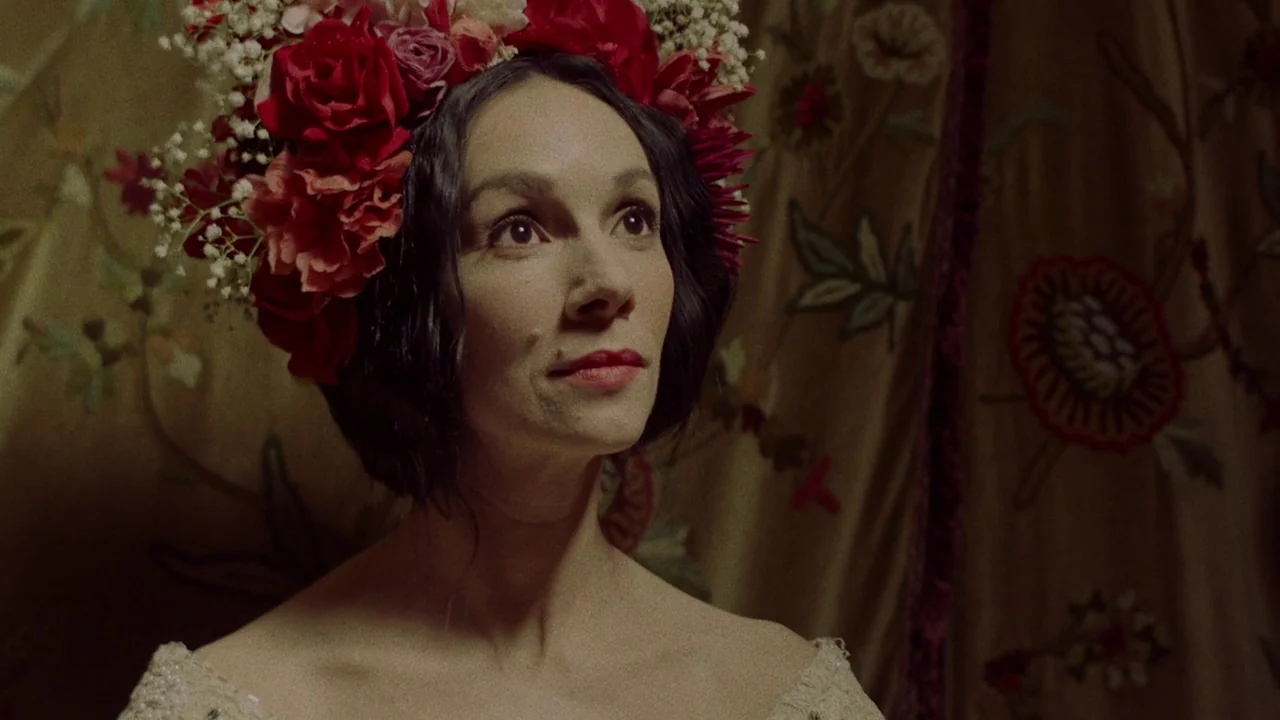 The Field Guide to Evil (2018) movie still: closeup — A woman wears a large crown of fresh red roses and baby's breath. She sits before an ornate…