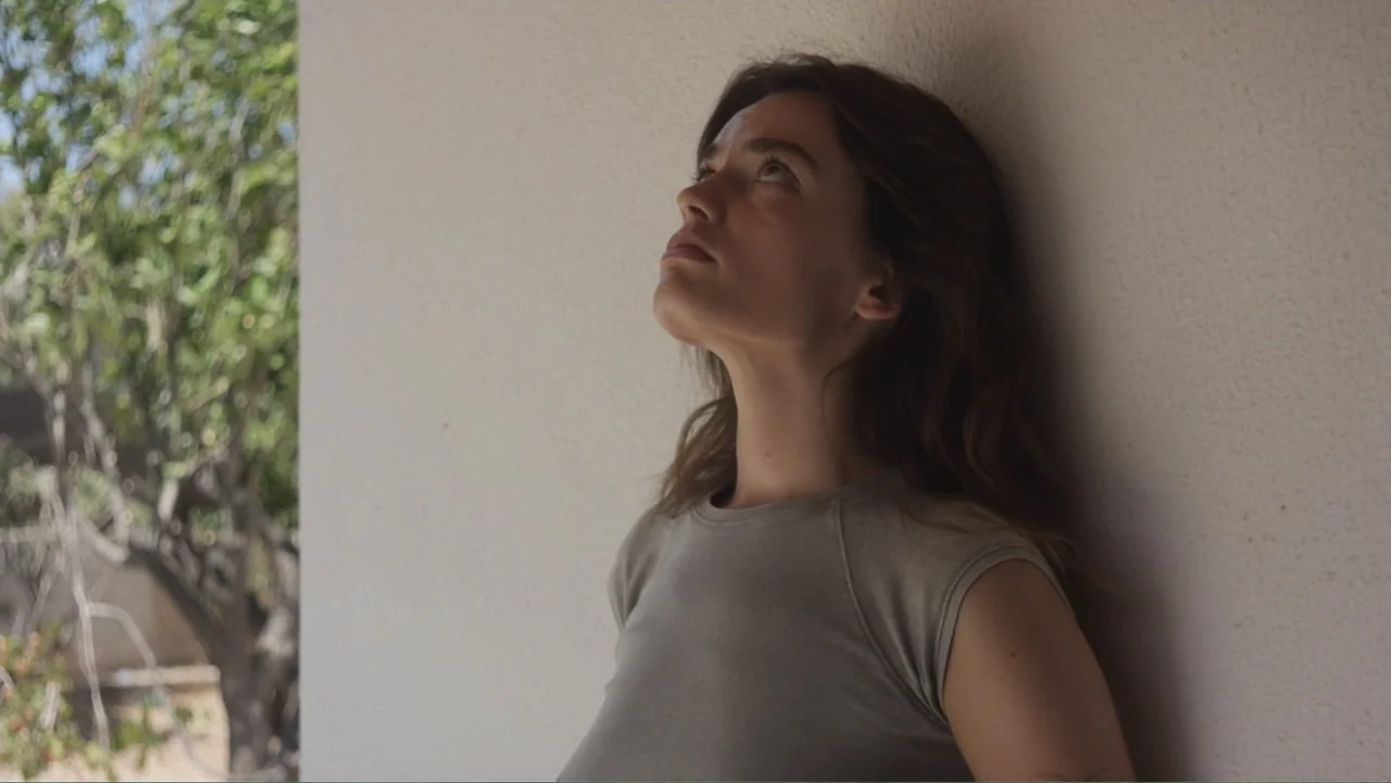 Hot Milk (2025) movie still: medium — A woman leans against a textured white wall in an outdoor shaded area. She gazes upwards towards the light…