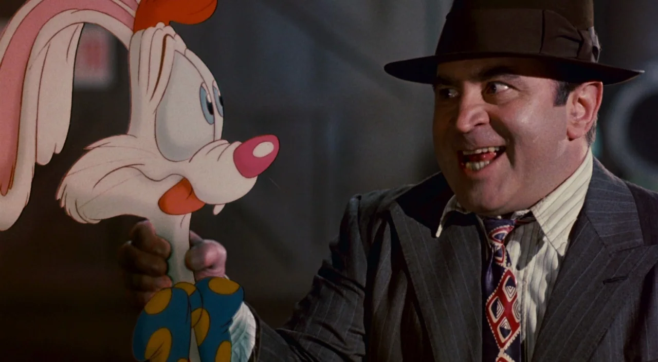 Who Framed Roger Rabbit (1988) movie still: medium — A dimly lit room with muted walls provides a backdrop for a man holding a cartoon. The man…