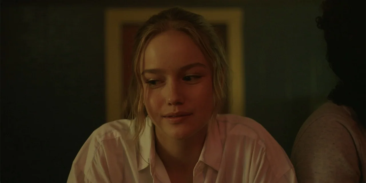 La coda del diavolo (2024) movie still: closeup — A dimly lit interior space features dark walls behind a seated subject. A young woman in a white…