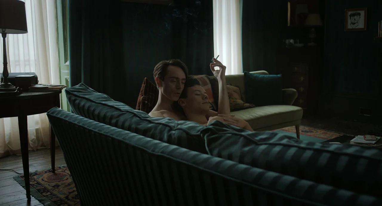 My Policeman (2022) movie still: medium — A dimly lit room with teal walls and dark curtains features two sofas. Two men without shirts lean back…