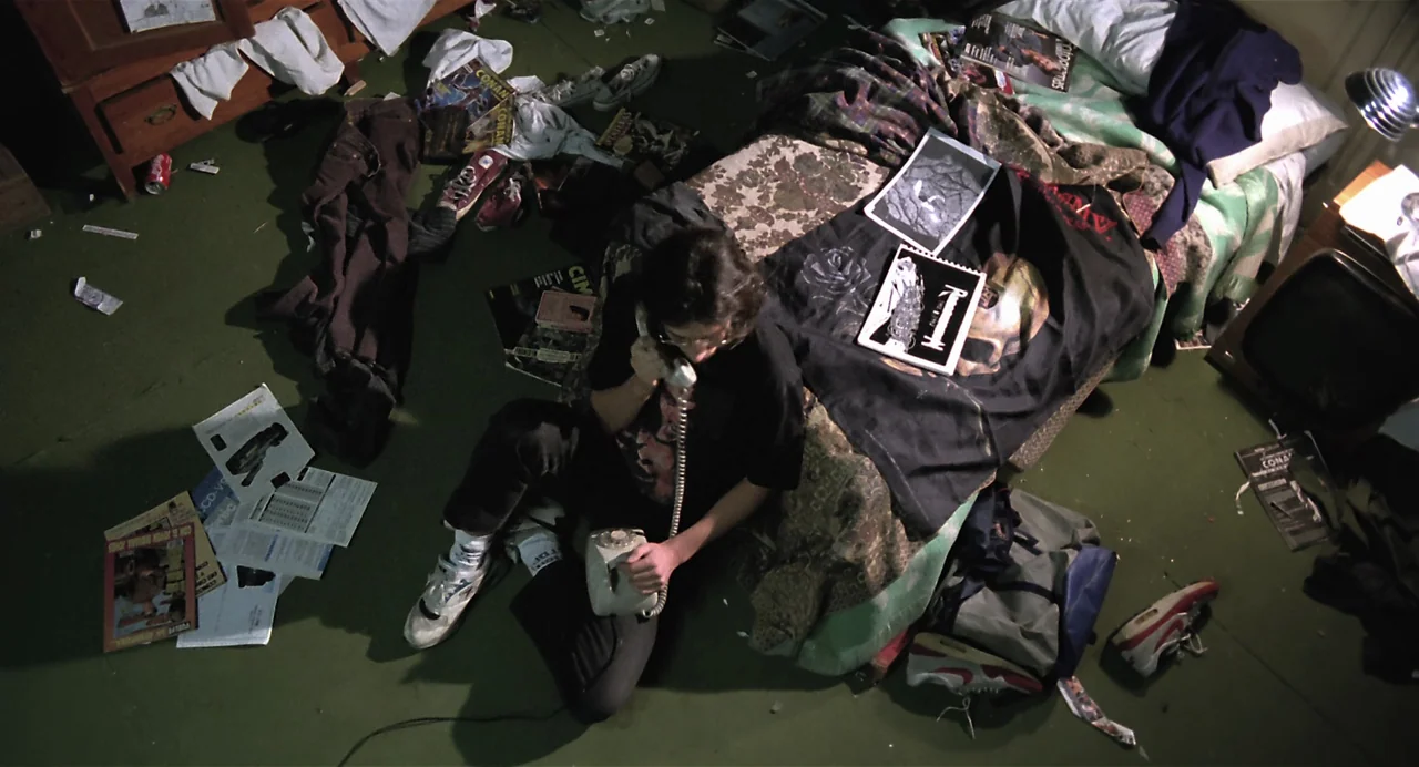 Thesis (1996) movie still: wide — A messy, cluttered space features a green carpet covered in magazines, shoes, and clothing. A person sits on the…