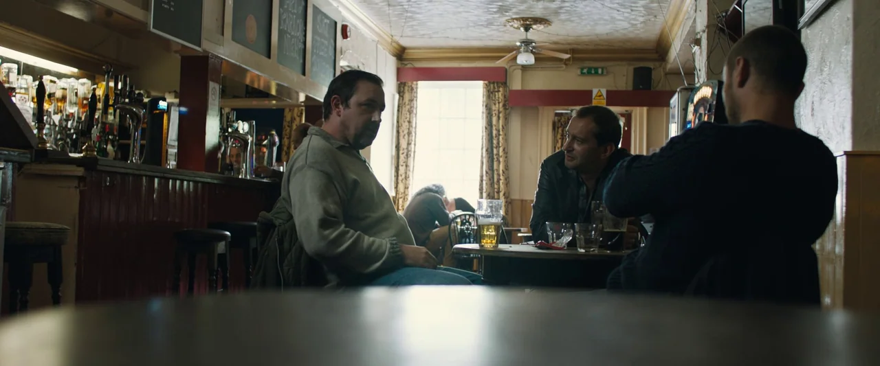 Black Sea (2014) movie still: medium — A dimly lit traditional pub interior features wooden fixtures and a rear window source. Three men occupy a…