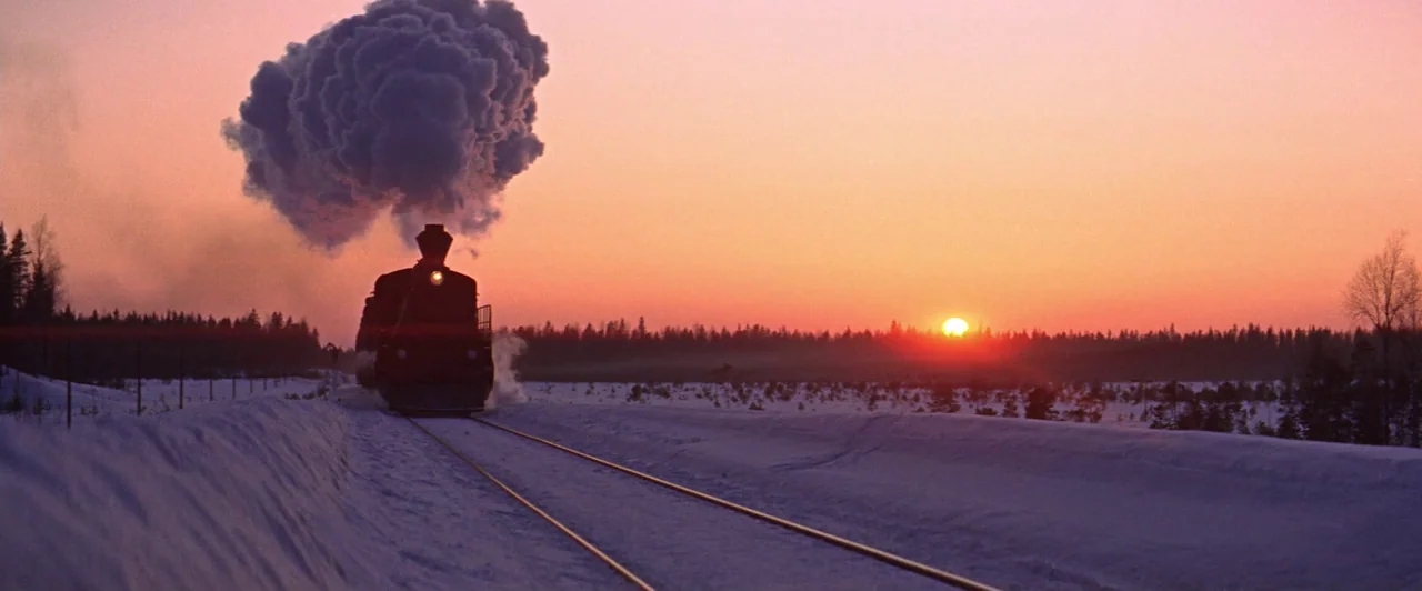 Doctor Zhivago (1965) movie still: wide — A steam locomotive approaches through a snowy, flat landscape during a vivid sunset. The train moves along…