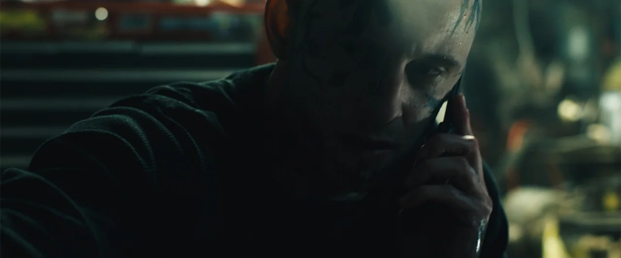 Skin (2009) movie still: closeup — A dimly lit room contains dark metallic surfaces and tools in the background. A man with facial tattoos holds a…
