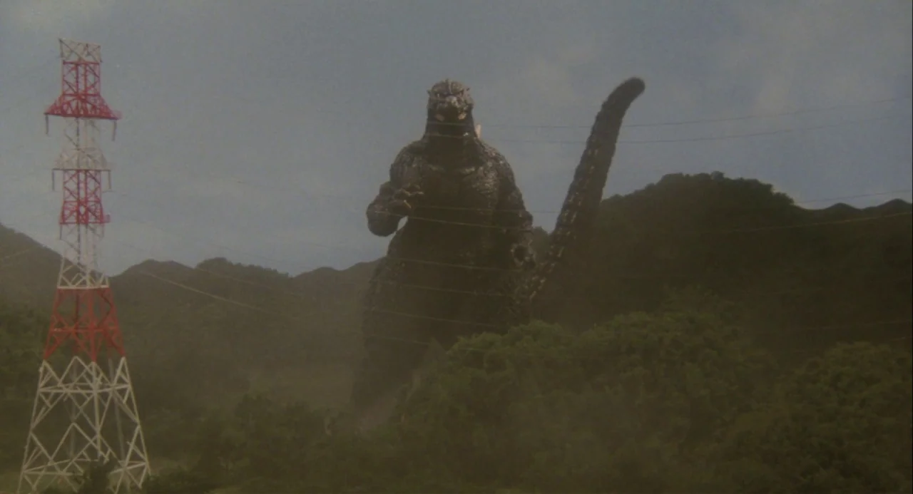 Godzilla vs. Mechagodzilla II (1993) movie still: wide — A gargantuan monster stands amongst green rolling hills and dense vegetation under a cloudy…