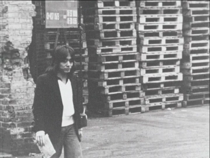 Catalogue of Memory (1977) movie still: fullbody — A man walks past high stacks of wooden crates beside a weathered brick wall. He carries a…