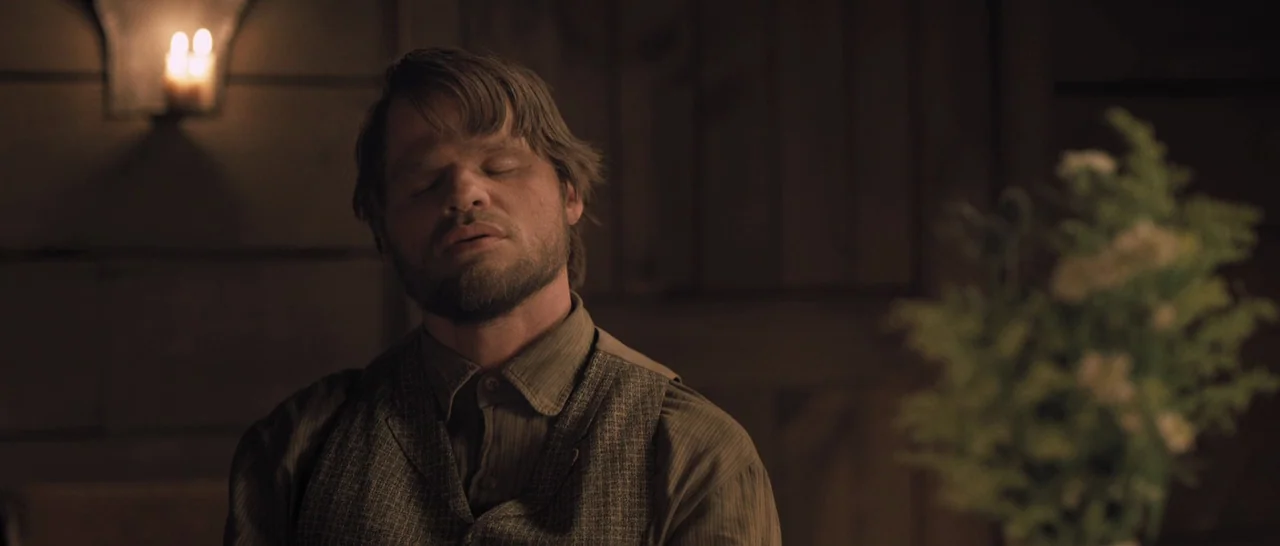 The Homesman (2014) movie still: medium — A dimly lit room features a man sitting in front of wooden paneling. A floral bouquet is out of focus in…