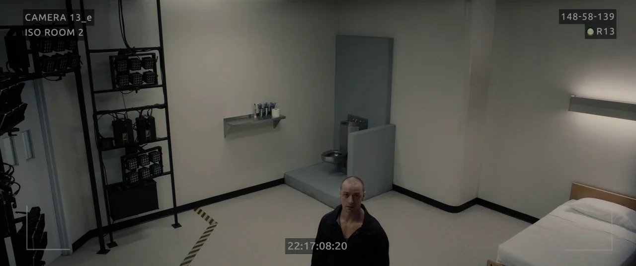 Glass (2019) movie still: wide — A stark white detention room features an integrated metal toilet and surveillance equipment. A solitary man in a…