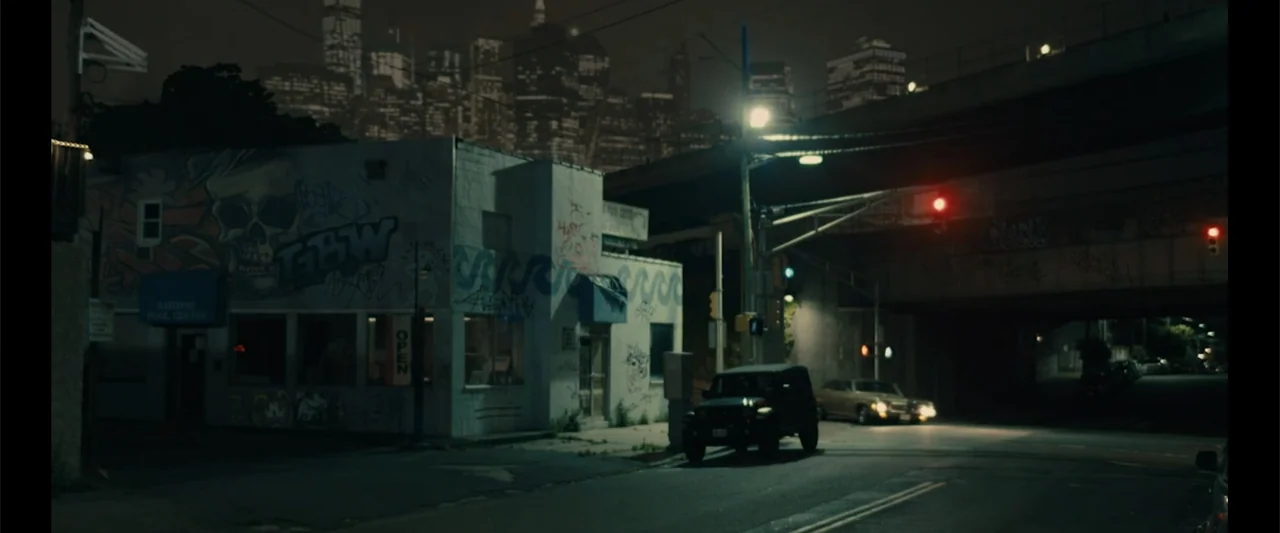 Howl (2015) movie still: wide — An urban intersection at night features a graffiti-covered building under a highway overpass. A dark SUV and a…