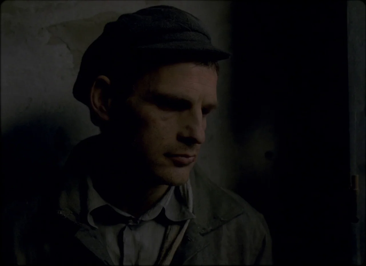Son of Saul (2015) movie still: closeup — A dimly lit, shadowed space provides a stark backdrop for the close-up portrait. A man wearing a flat cap…
