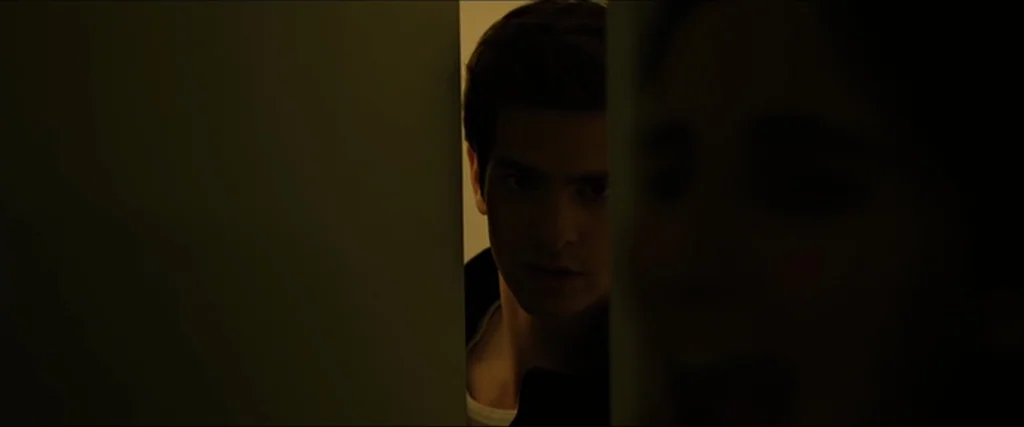 The Social Network movie still: closeup — A dark space with a vertical gap revealing a illuminated interior room. A young man stares intently…