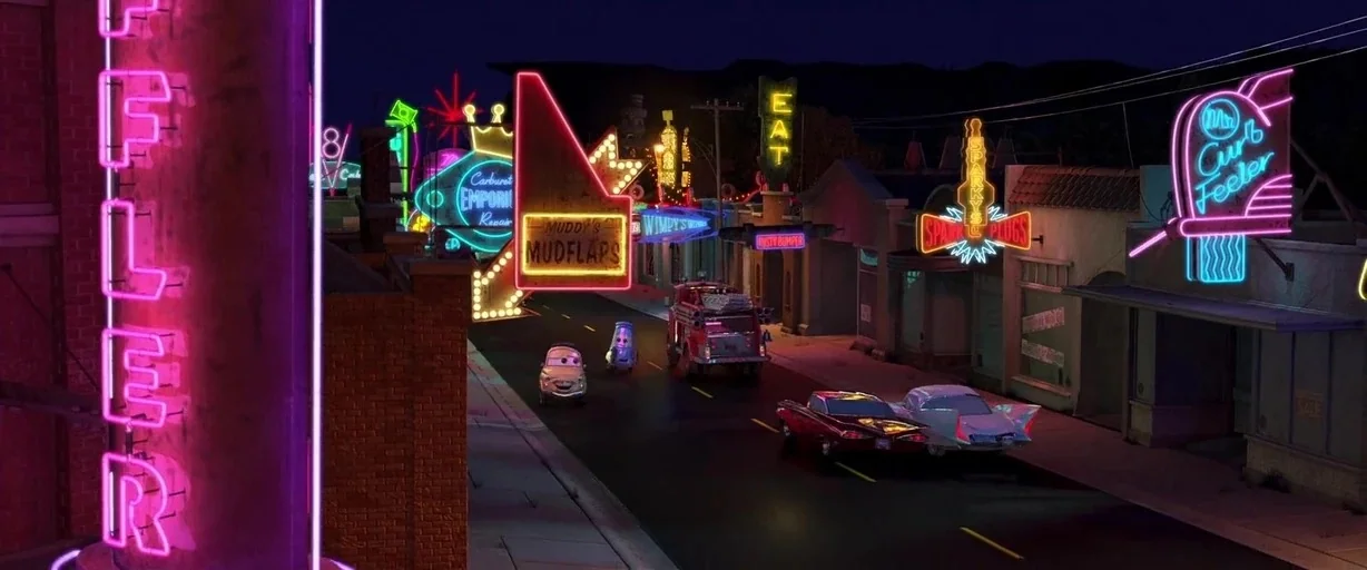 Cars (2006) movie still: wide — A nighttime main street is illuminated by numerous glowing neon signs above small buildings. Several animated cars…