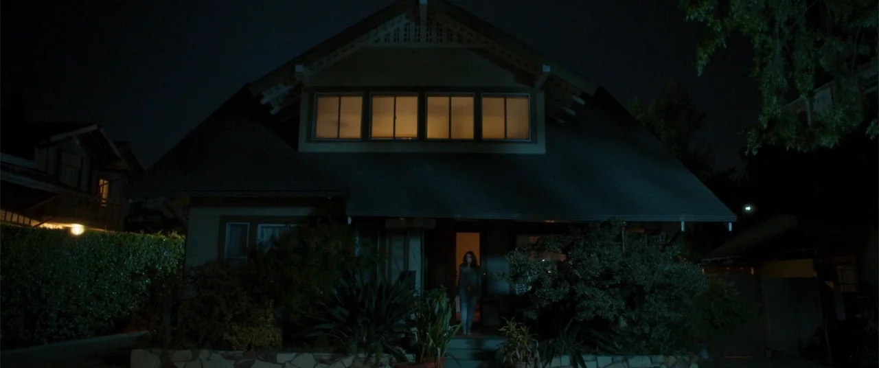 Phobias (2021) movie still: wide — A dark two-story suburban home at night features illuminated windows and a dimly lit doorway. A lone woman stands…