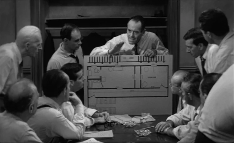 12 Angry Men (1957) movie still: medium — A group of men gather around a large table in a sparsely furnished room. One man stands and points at a…