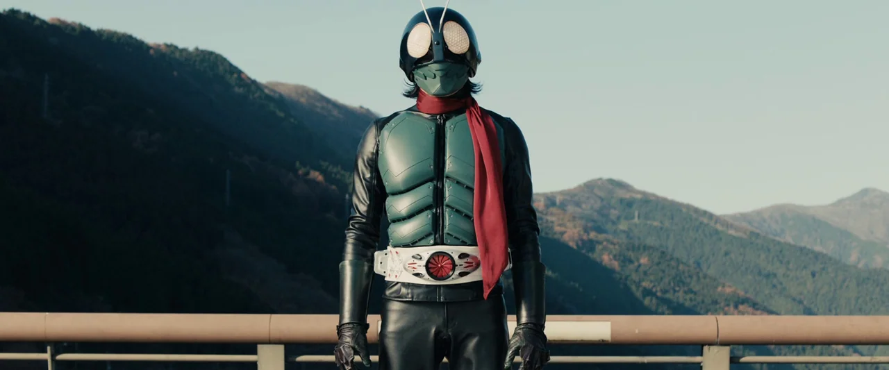Shin Kamen Rider (2023) movie still: fullbody — A figure in a green and black insect-themed costume stands on a road barrier. Behind the subject are…