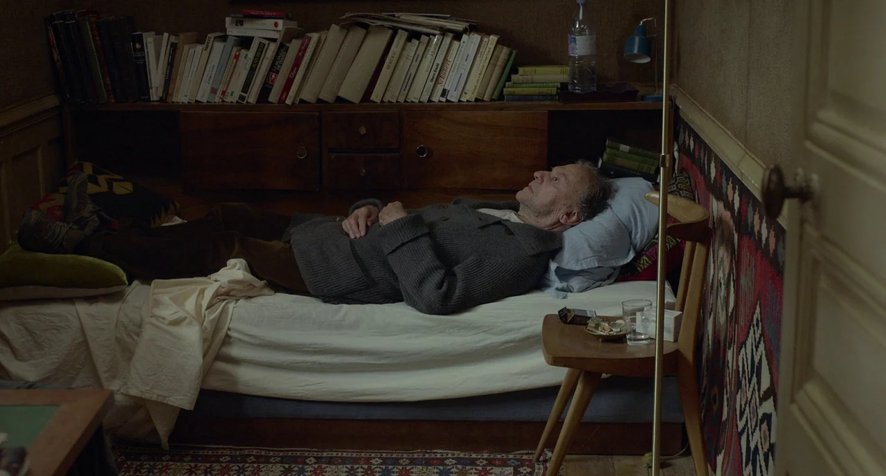 Amour (2012) movie still: medium — A small cluttered space contains a single bed positioned against a wall of books. An elderly man rests on the bed…