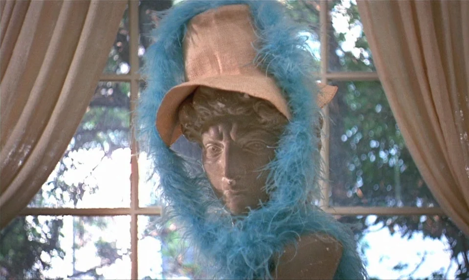 Lions Love (1969) movie still: closeup — A stone sculpture head sits in front of a window with beige curtains. The sculpture wears a straw-colored…
