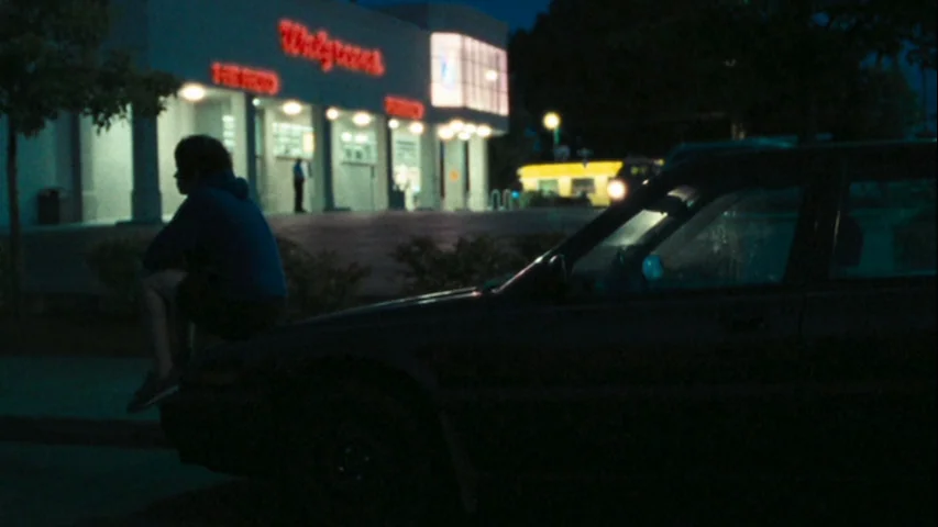 Wendy and Lucy (2008) movie still: wide — A dark, quiet parking lot at night features a Walgreens store illuminated by neon. A young man sits…