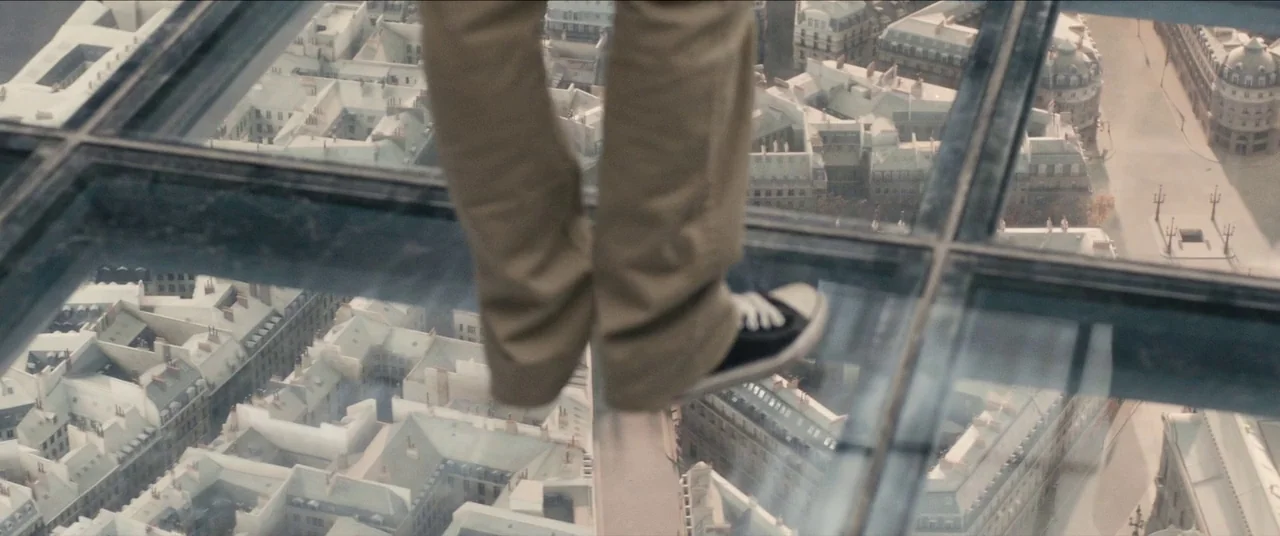 Simon Killer movie still: closeup — A glass floor pane looks down upon a dense grid of urban Parisian buildings. The lower legs of a person wearing…