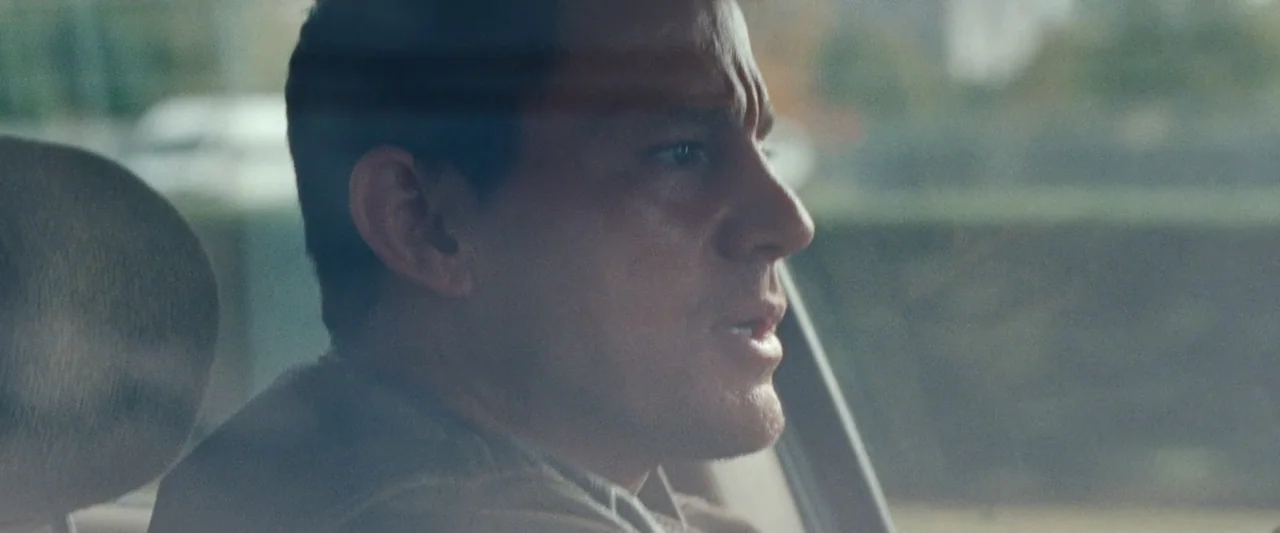 Roofman (2025) movie still: closeup — The interior of a moving vehicle during the day with sunlight streaming inside. A man in his thirties sits…