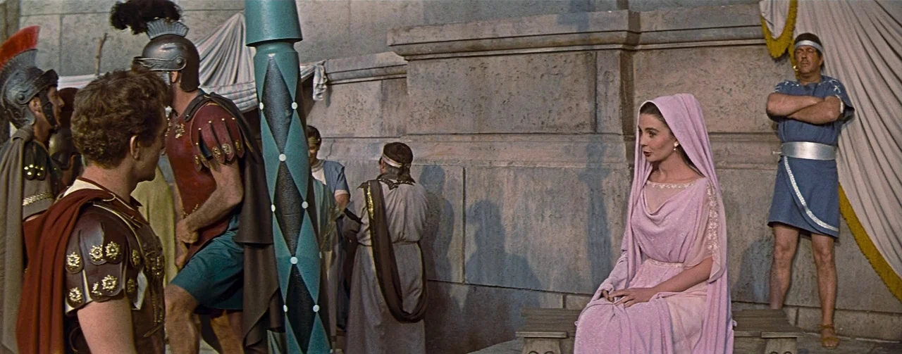 The Robe (1953) movie still: wide — A stone courtyard features ancient architectural elements and draped cloth hangings. A woman sits on a bench…